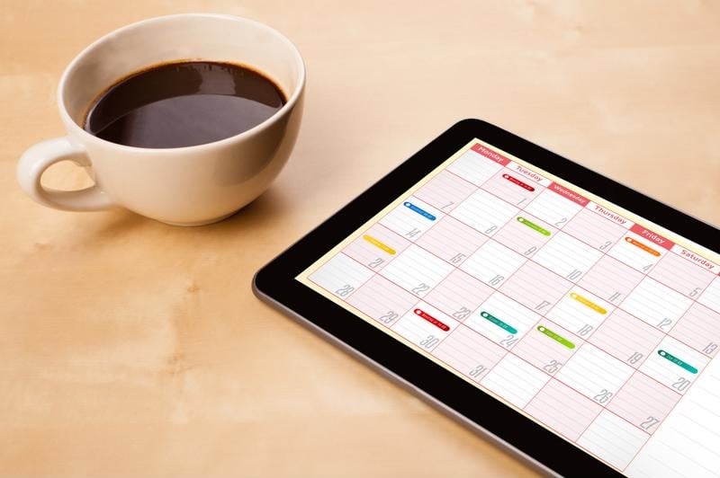 Calendar with dates blocked off in different colors on a tablet sitting on a table next to a cup of coffee.