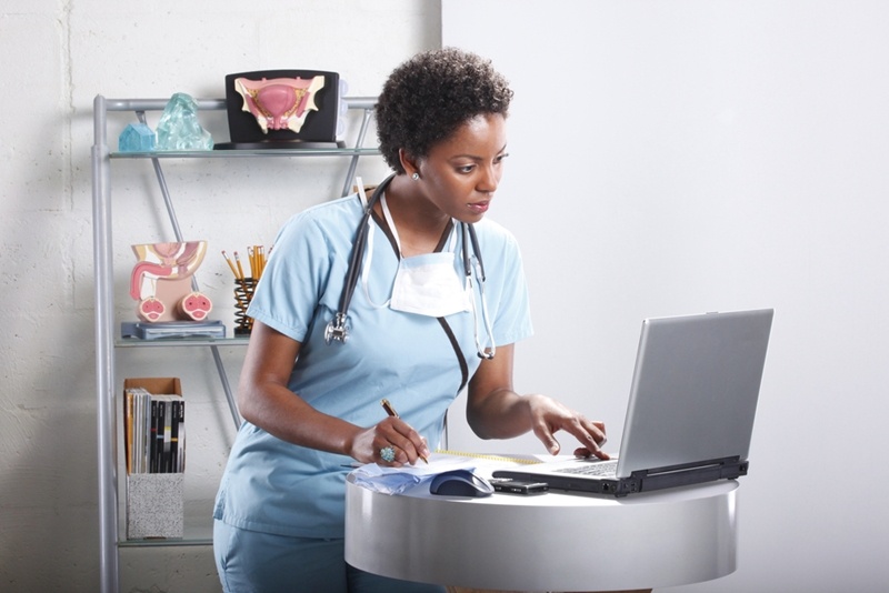 Young doctor sitting in her office with stethoscope around her neck working on laptop computer with book nearby. 