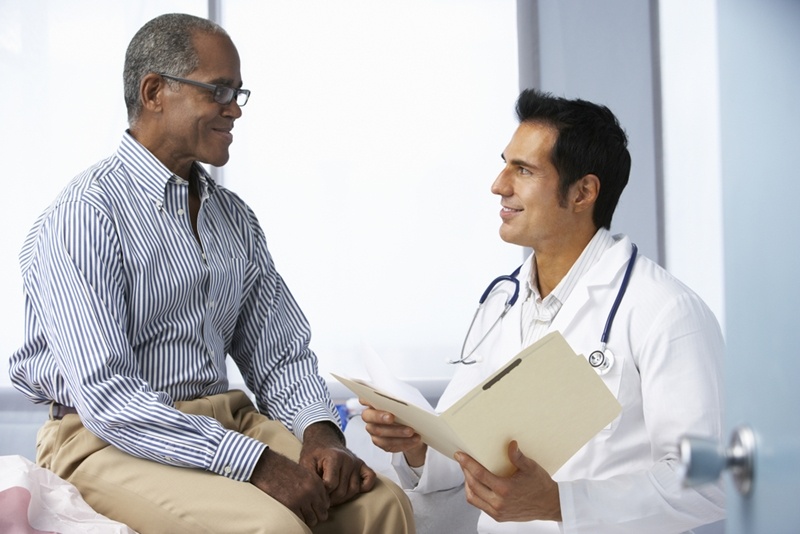 Younger male doctor holding open folder sitting with older, male patient. 