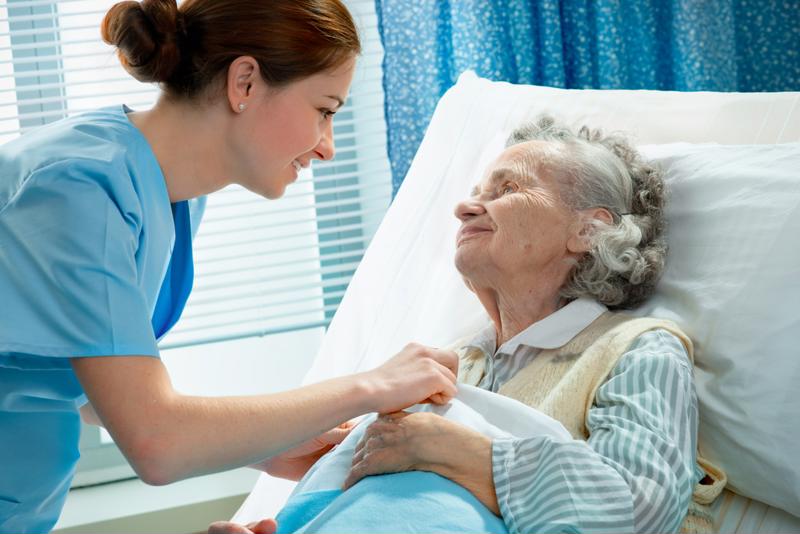Young nurse leaning over the bed and fixing the blankets of a smiling, elderly patient. 