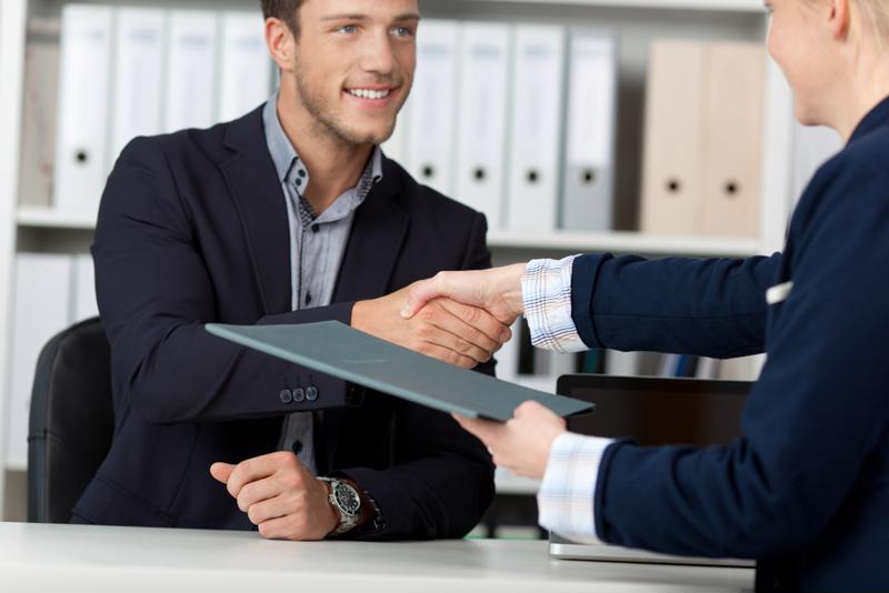 Job interview - one man in a suit shaking the hand of another man wearing a suit and holding a folder. 
