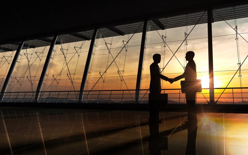 Two people shaking hands in a long hallway with windows at sunset. 